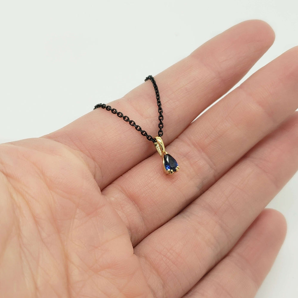 Pear blue sapphire pendant in yellow gold on black chain, shown in hand for scale
