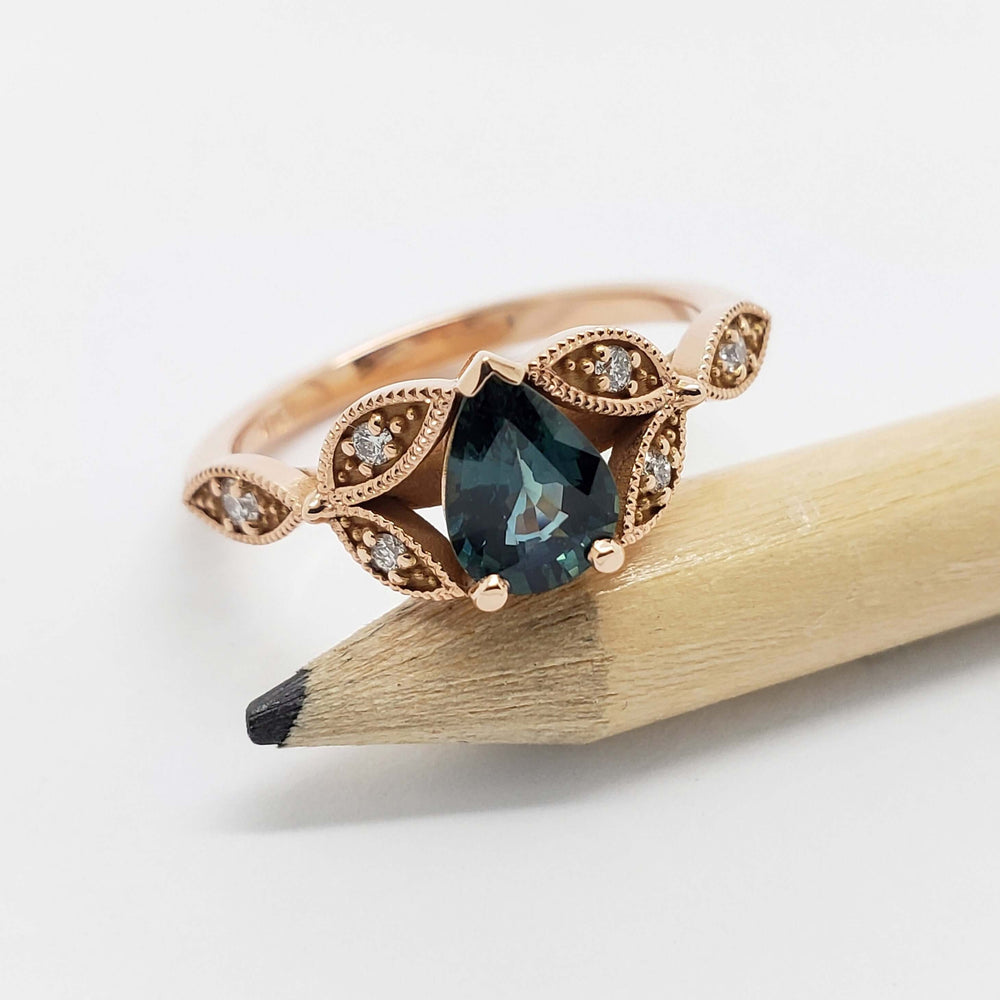Pear teal sapphire and diamond leaf ring in 14k rose gold, displayed on a pencil
