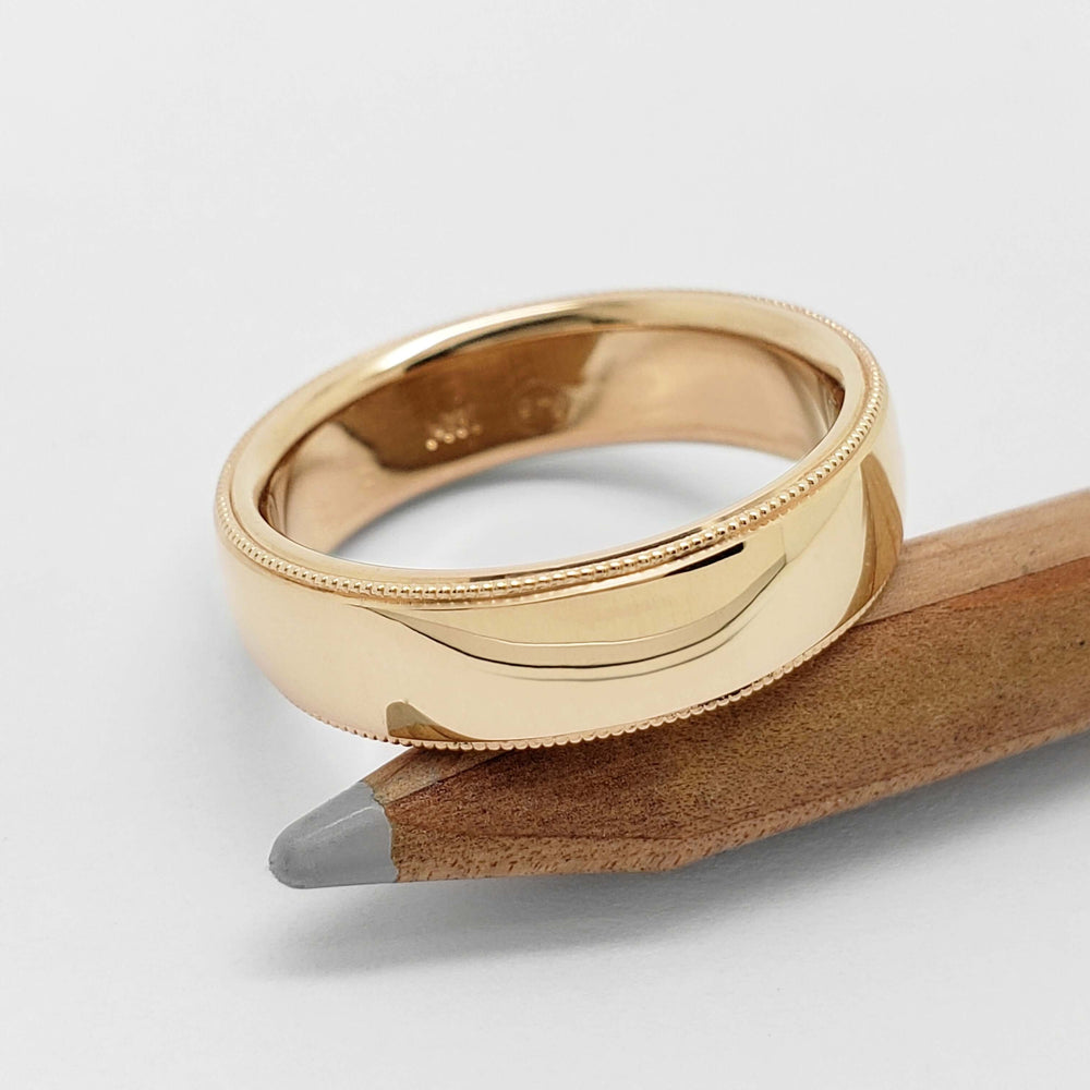 Polished 14kt yellow gold wedding ring with millgrain edge resting on a pencil