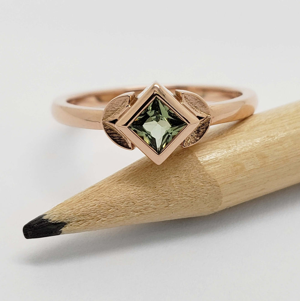Princess cut green sapphire ring in rose gold with leaf accents, resting on a pencil