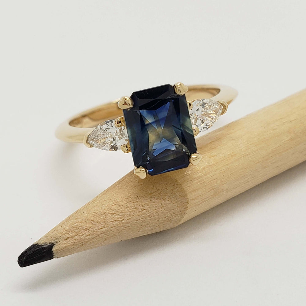 Radiant cut blue sapphire and pear lab diamond ring in yellow gold, displayed on a pencil