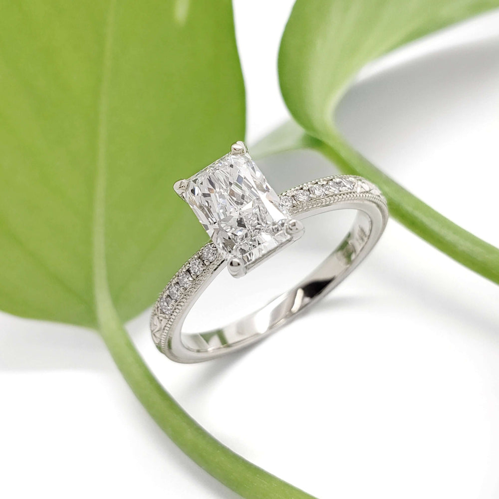 Radiant cut lab diamond engagement ring in 14kt white gold with pave band, beside green leaf