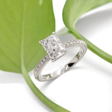Radiant cut lab diamond engagement ring in 14kt white gold with pave band, beside green leaf