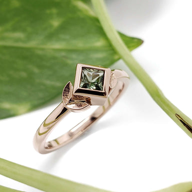 Rose gold engagement ring with green princess cut sapphire and leaf motif, on white background with green leaves