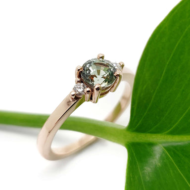 Rose gold engagement ring with green sapphire and diamonds, displayed on a green leaf
