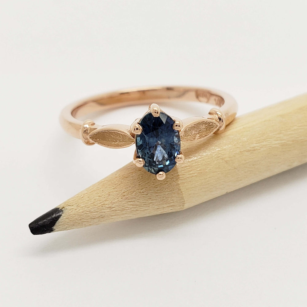 Rose gold ring with blue Montana sapphire and leaf motif, displayed on a pencil