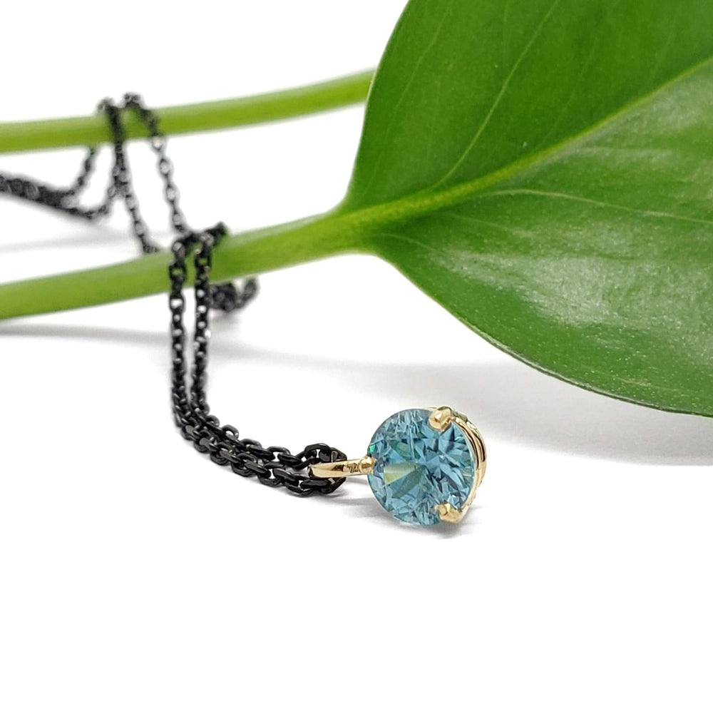 Round blue zircon pendant in 14kt yellow gold on black chain, styled with green leaf