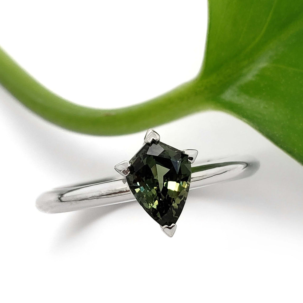 Shield cut green sapphire engagement ring in white gold beside a green leaf