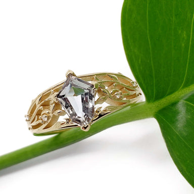 Shield cut sapphire and diamond filigree engagement ring in yellow gold resting on a green leaf