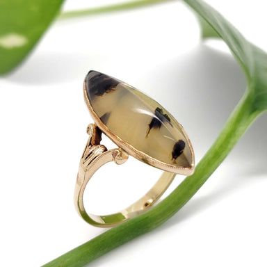 Side angled view of marquise dendritic agate set in detailed 10kt yellow gold ring against a leaf
