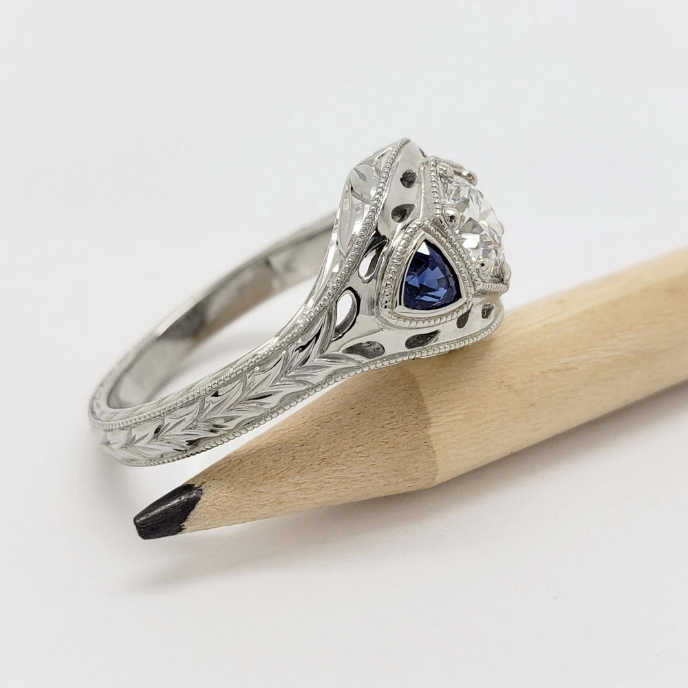 Side view of engraved white gold ring with blue sapphire accent and lab diamond, next to a pencil