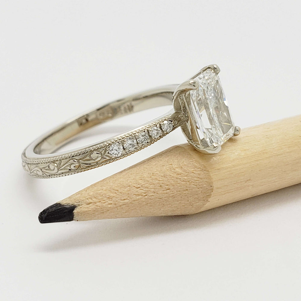 Side view of engraved 14kt white gold engagement ring with radiant lab diamond and pave-set band, next to pencil