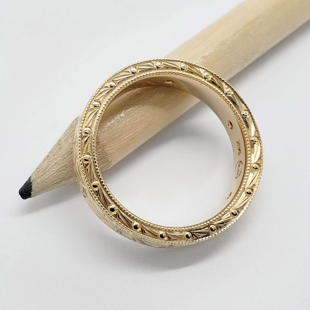 Side view of engraved 14kt yellow gold wedding ring with millgrain and bead details, on pencil