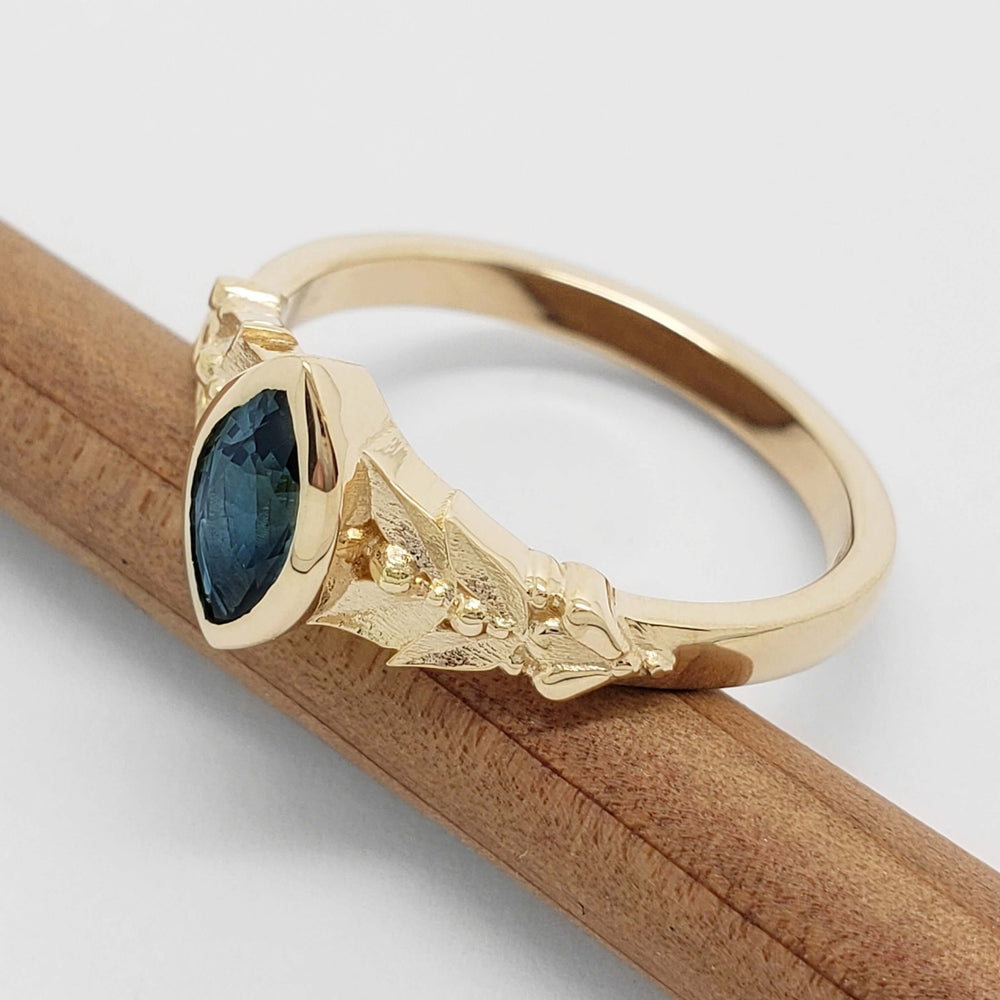 Side view of 14kt yellow gold ring with marquise blue sapphire and engraved leafy band