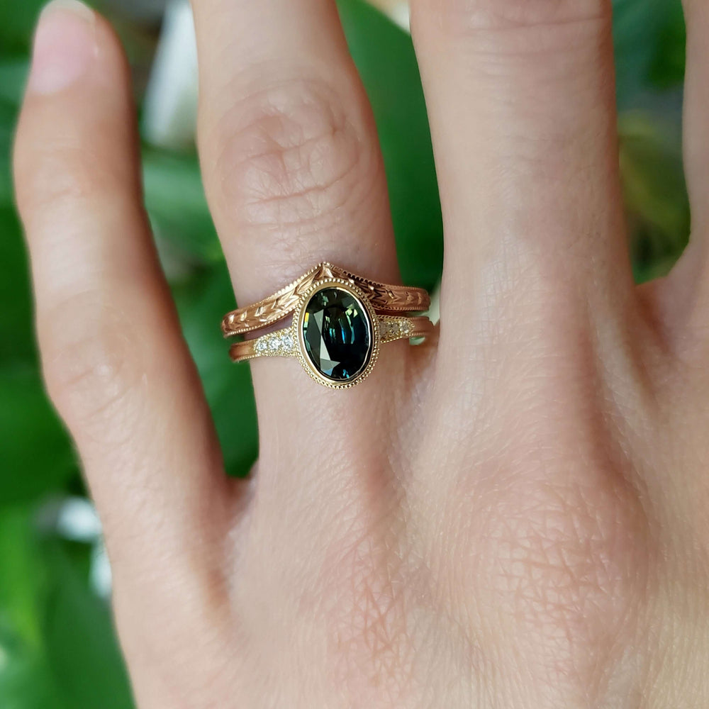Teal sapphire and diamond engagement ring stacked with engraved yellow gold band on hand