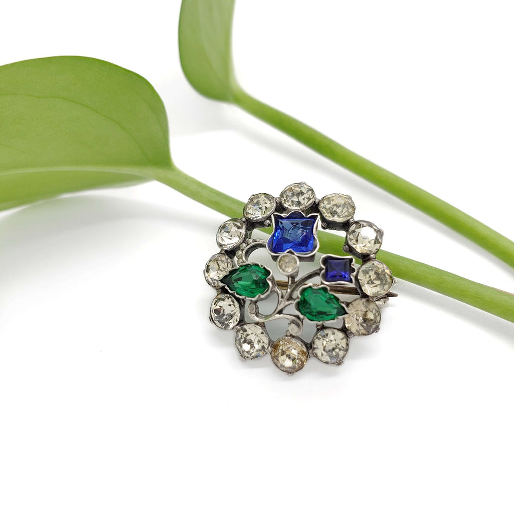 Sterling silver floral brooch with blue and green glass stones displayed on a green leaf