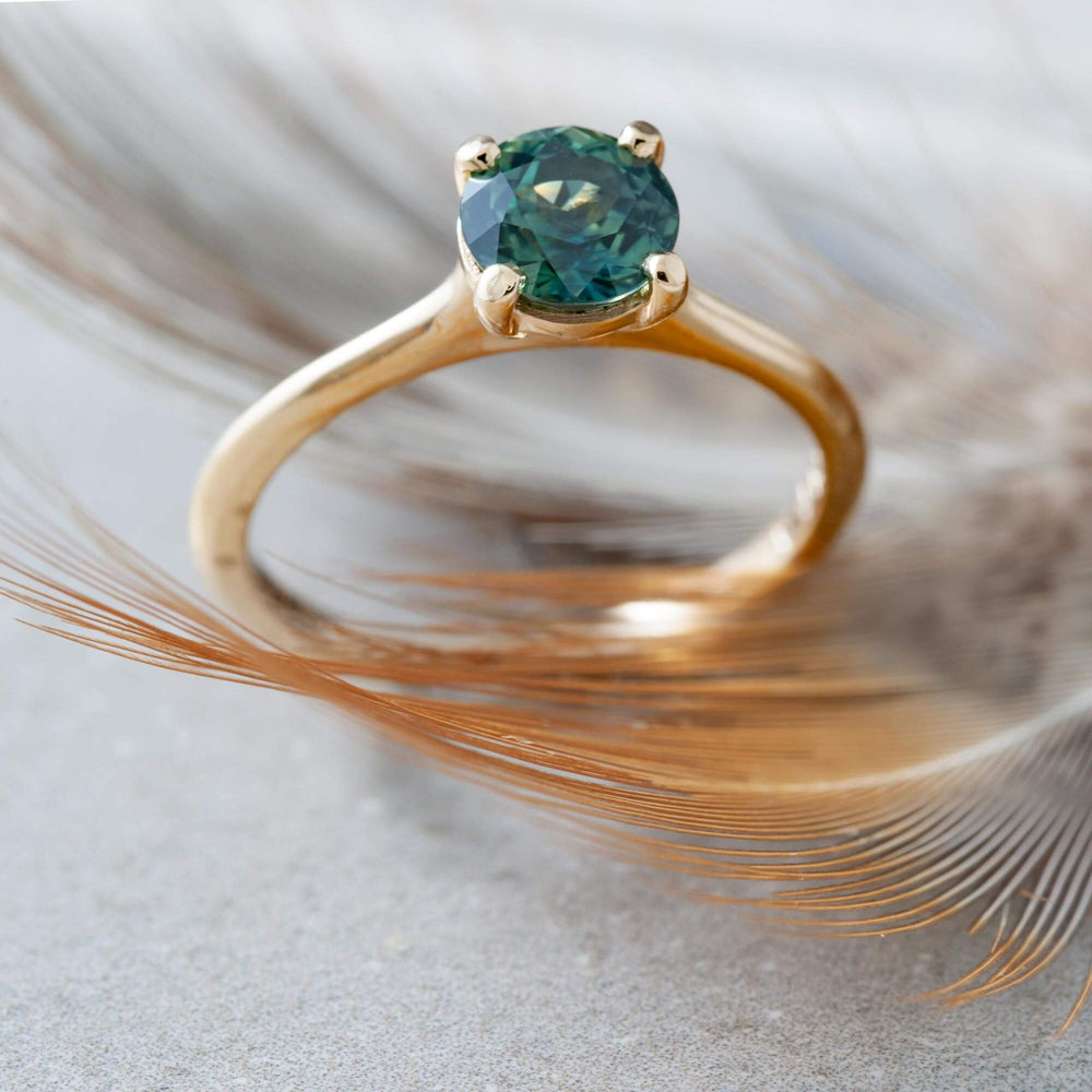 Teal Australian sapphire engagement ring in yellow gold resting on a feather