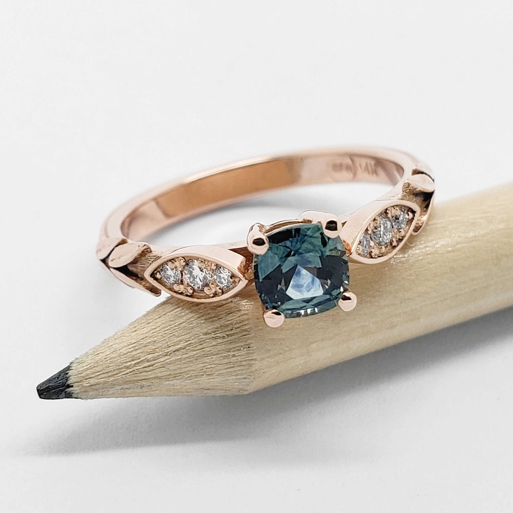 Cushion cut teal sapphire and diamond rose gold ring displayed on a pencil