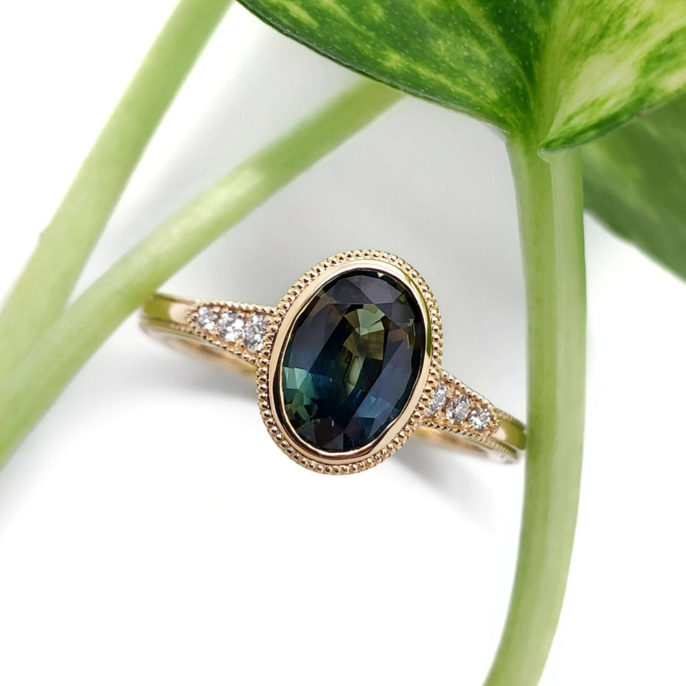 Teal Australian sapphire and diamond engagement ring in yellow gold, shown with green plant
