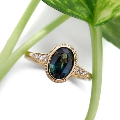 Teal Australian sapphire and diamond engagement ring in yellow gold, shown with green plant