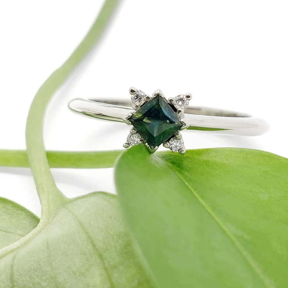 Teal sapphire and lab diamond white gold ring resting on a green leaf