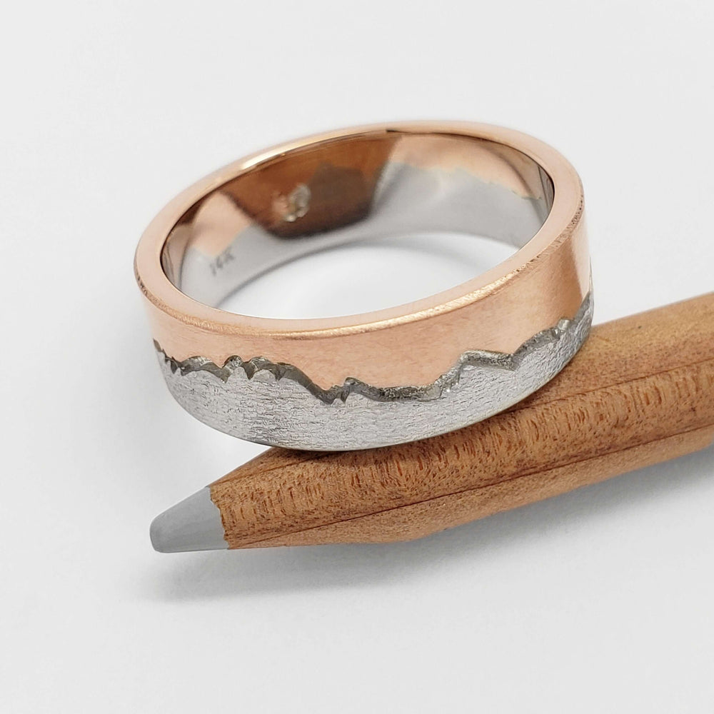 Textured 14k white and rose gold mountain range wedding band resting on a pencil