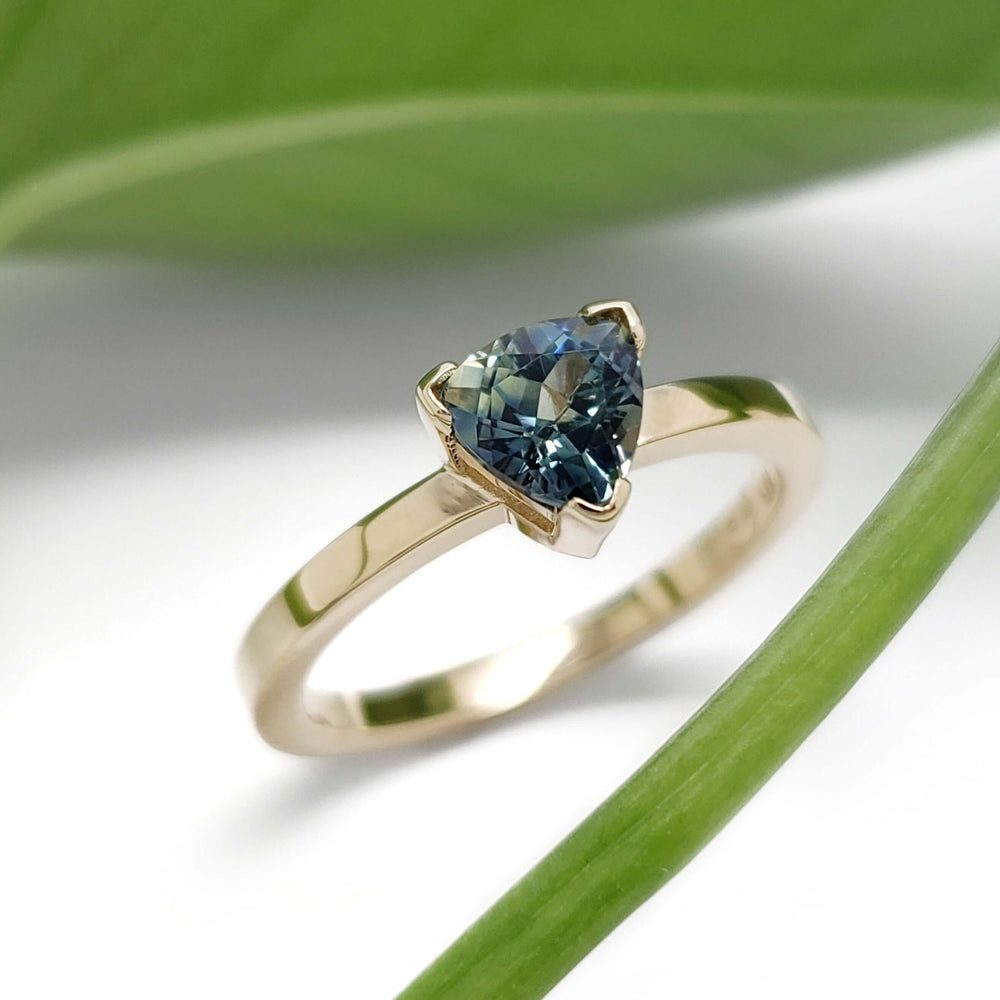 Trillion cut teal Australian sapphire ring in yellow gold, displayed with green leaf