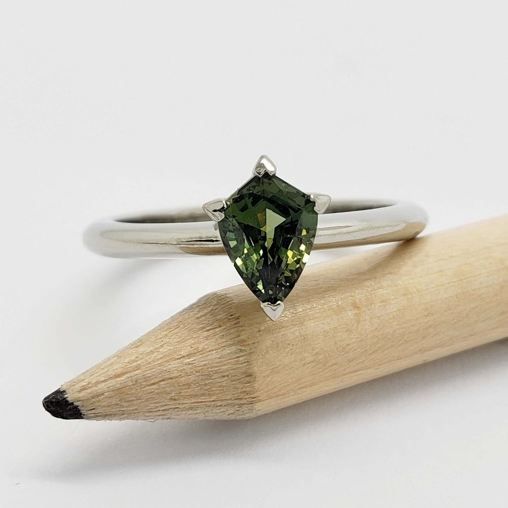 White gold engagement ring with shield cut green sapphire resting on a pencil
