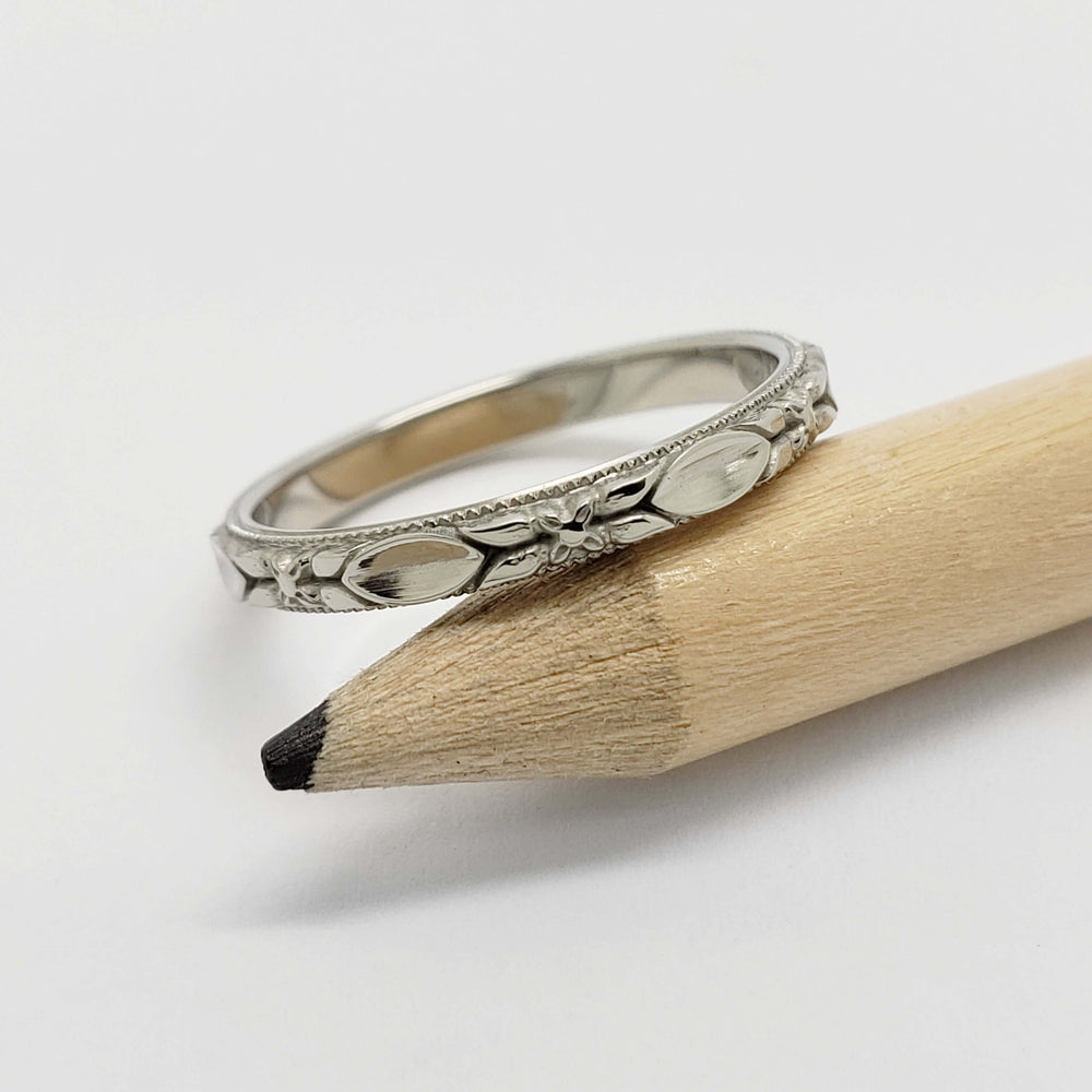 White gold wedding ring with engraved floral pattern shown on a pencil for scale