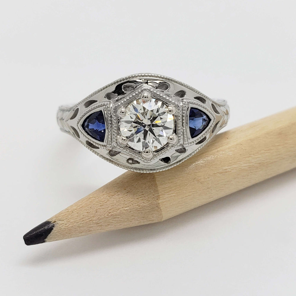 White gold engagement ring with round lab diamond and blue sapphires, displayed beside a pencil
