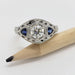 White gold engagement ring with round lab diamond and blue sapphires, displayed beside a pencil