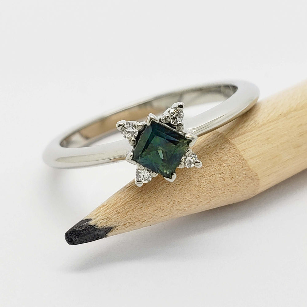 White gold ring with teal sapphire and lab diamonds displayed on a pencil
