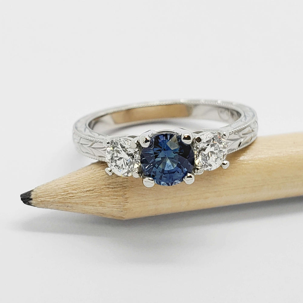 White gold three-stone ring with teal Montana sapphire and diamonds, shown on a pencil