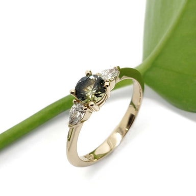 Handcrafted yellow gold ring with Montana sapphire and lab diamonds beside a green leaf