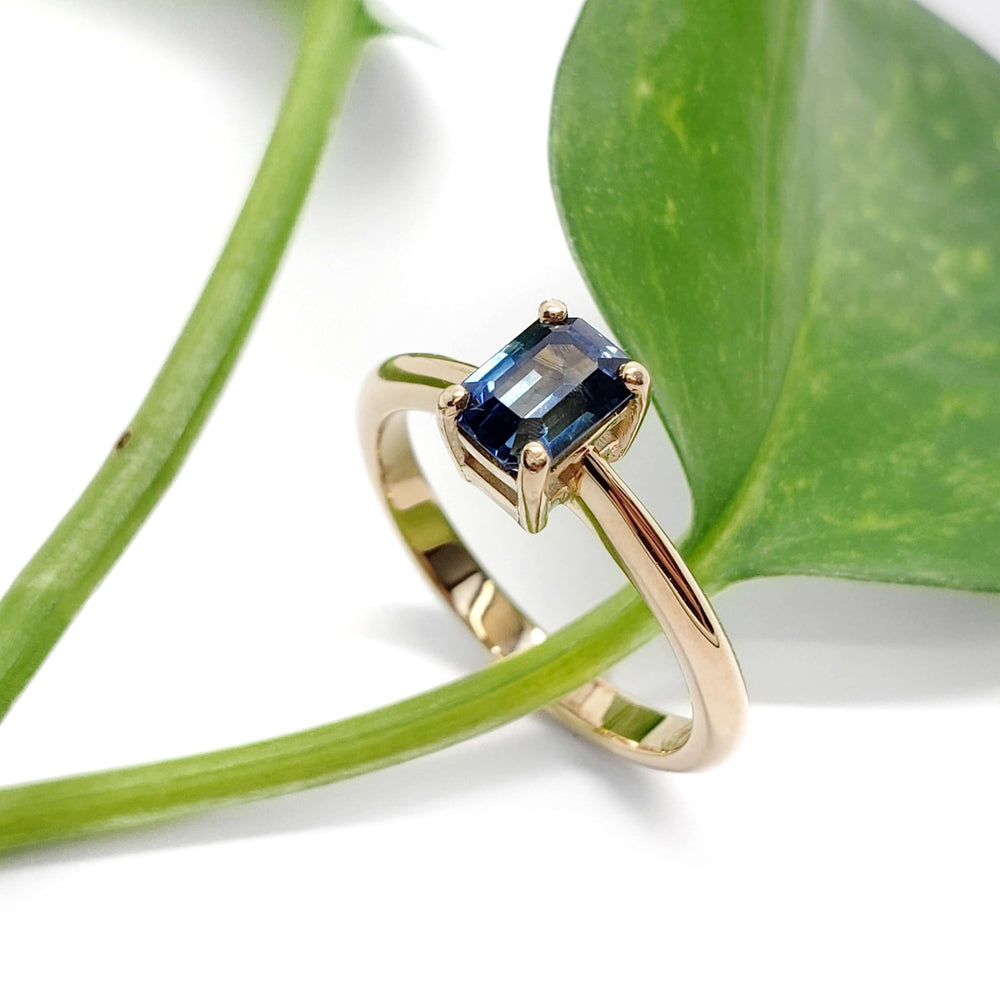 Yellow gold Montana sapphire ring with emerald cut blue stone beside green leaf