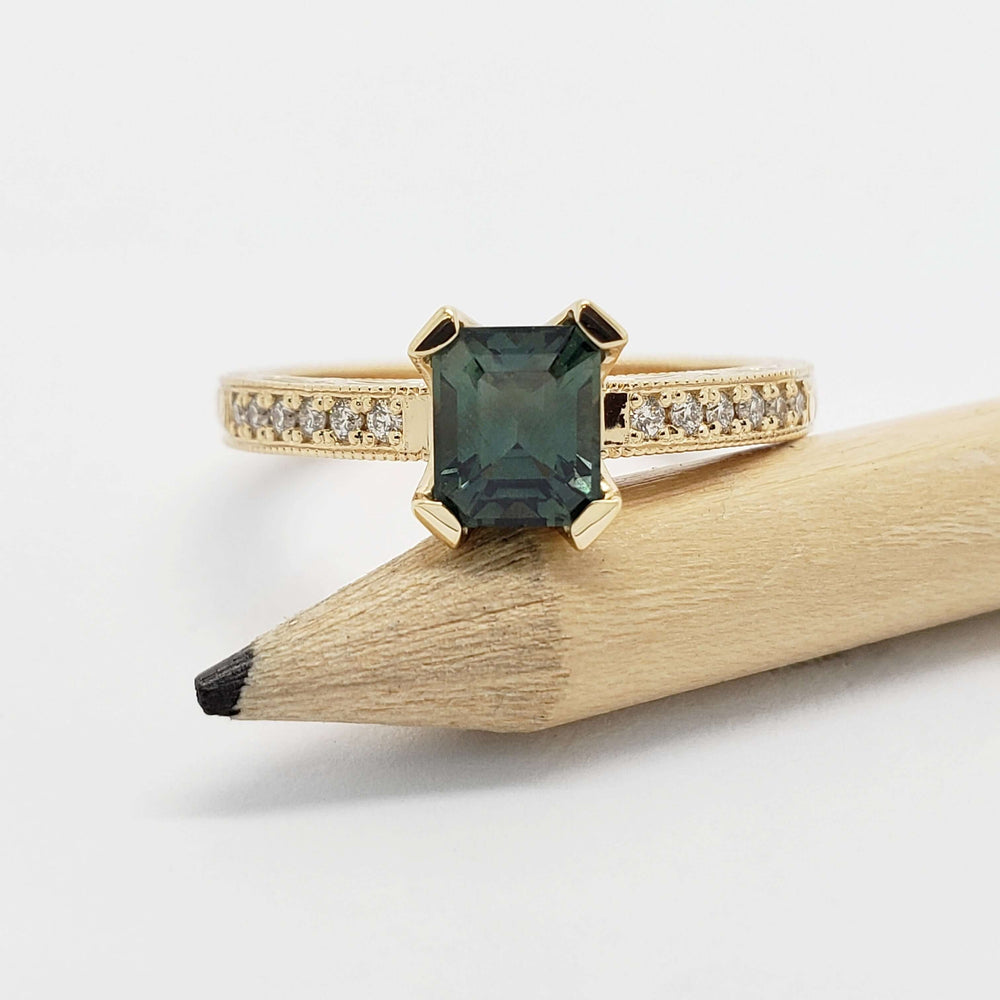 Front view of yellow gold engagement ring with emerald cut teal Australian sapphire and diamond band, beside pencil