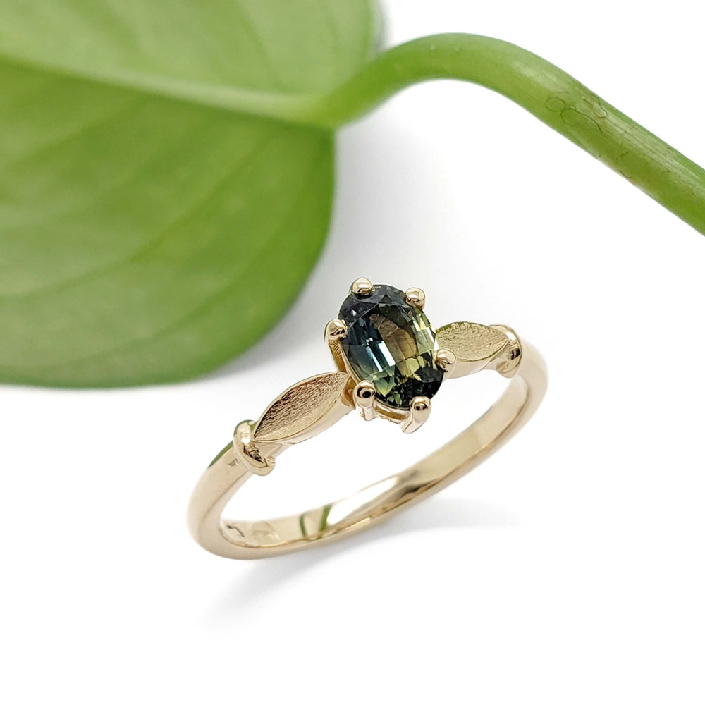Yellow gold engagement ring with oval blue-green Australian sapphire, leaf detail, beside green leaf