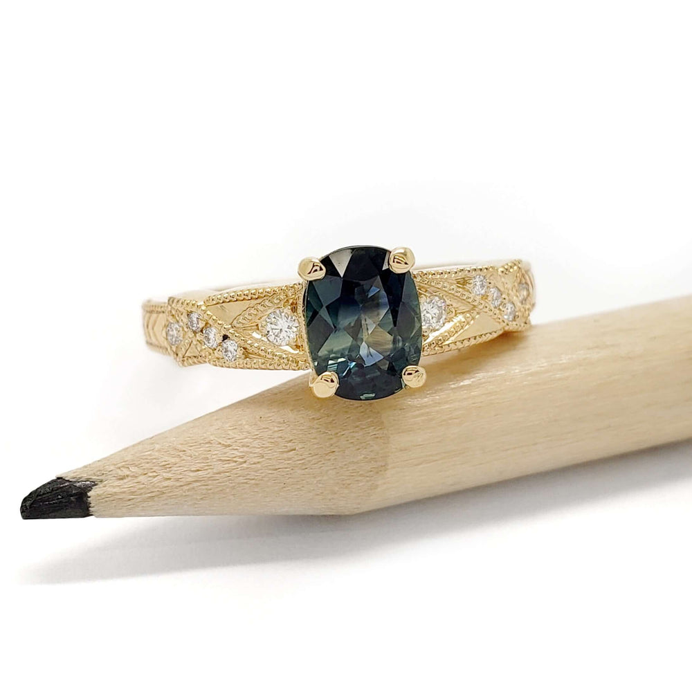 Yellow gold ring with blue-green sapphire and diamonds, displayed on a pencil