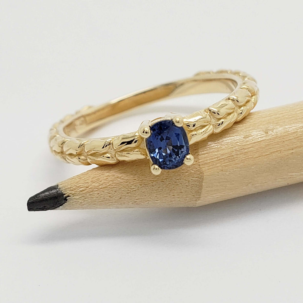 Yellow gold ring with blue sapphire and wheat pattern, displayed on a pencil
