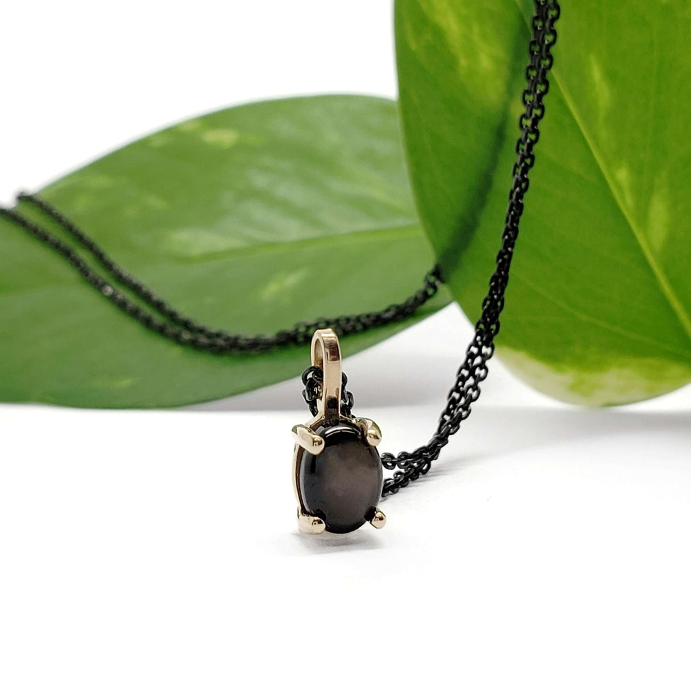 Star sapphire pendant in yellow gold with black chain, displayed against green leaves