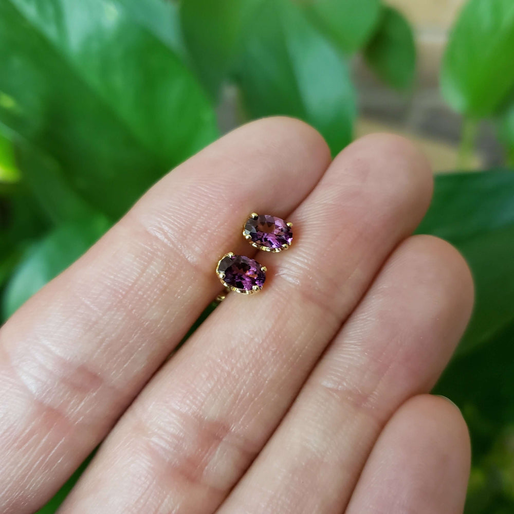 Hand holding 14kt yellow gold oval violet garnet stud earrings outdoors