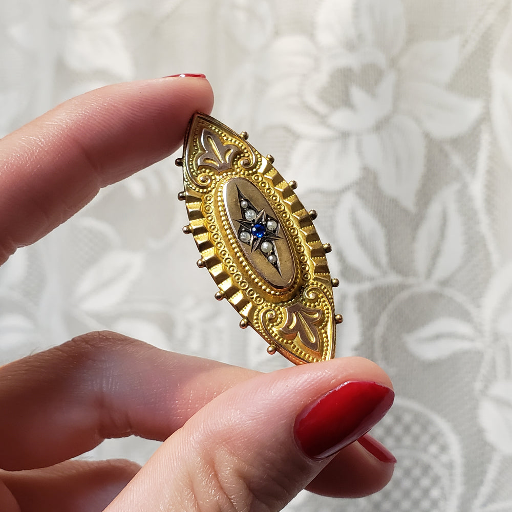 A hand with red painted fingernails holds a vintage gold brooch featuring an oval center with a blue and white floral motif, set against a white floral lace background.
