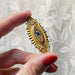 A hand with red painted fingernails holds a vintage gold brooch featuring an oval center with a blue and white floral motif, set against a white floral lace background.