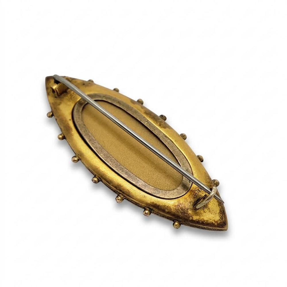 A vintage-style oval brooch with a distressed gold finish, featuring a pin-back mechanism with a silver-tone wire and small decorative studs along the border, shown on a plain white background.